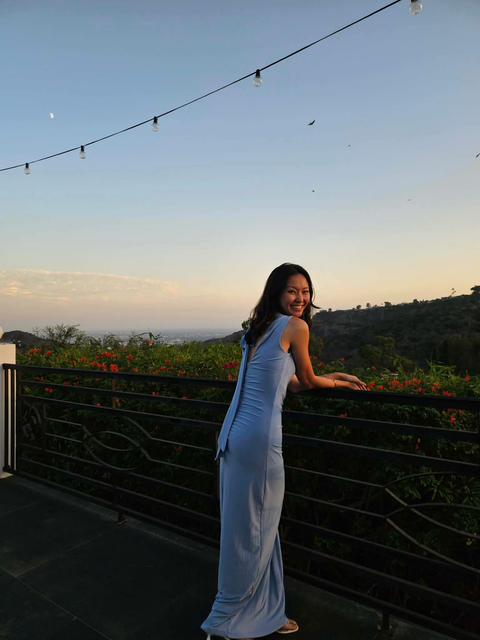 Melanie Renee in a long blue dress, smiling at a balcony railing at sunset